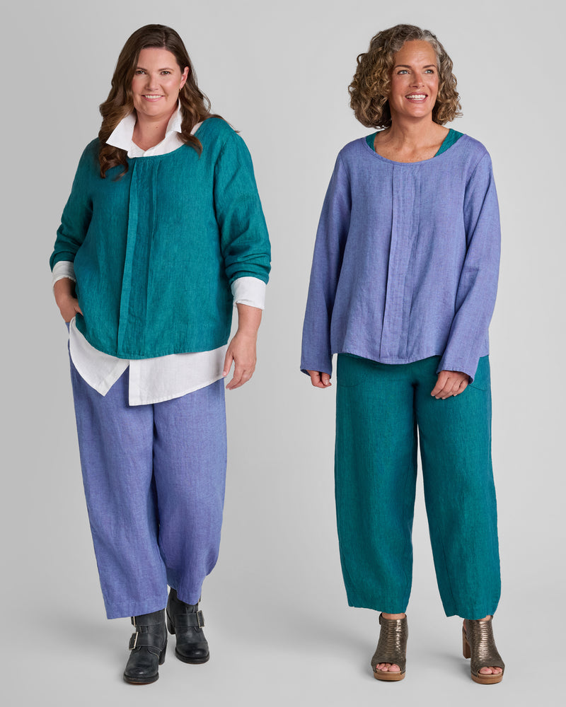 One woman in a blue linen shirt with purple linen pants and another woman with a purple linen shirt and blue linen pants